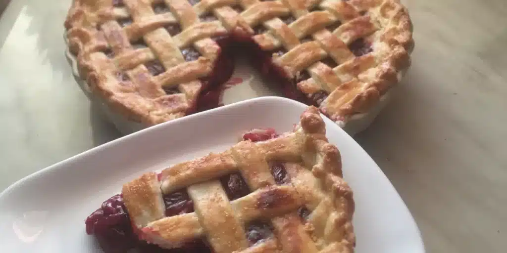 A baked round cherry pie with a lattice crust on top, displayed on a plate. A triangular piece has been cut out and is placed on a separate plate.