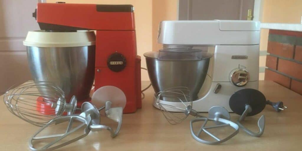 A white Kenwööd Chef and a vintage, red Kenwood Major stand mixer on a table, with a range of attachments.