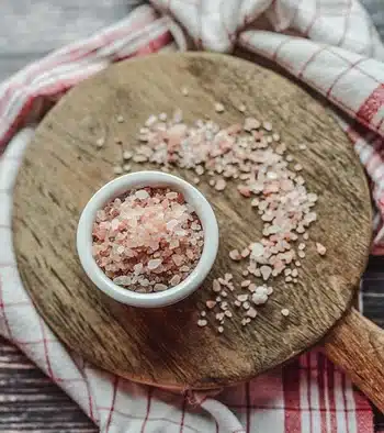 A bowl of pink Hymalayan salt on a wooden board also sprinkled with salt.